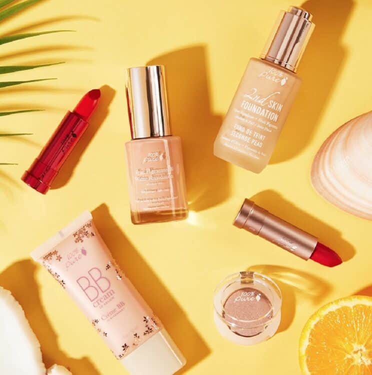 Three 100% Pure vegan foundations and two lipsticks on a yellow background accented with oranges, shells, and leaves.