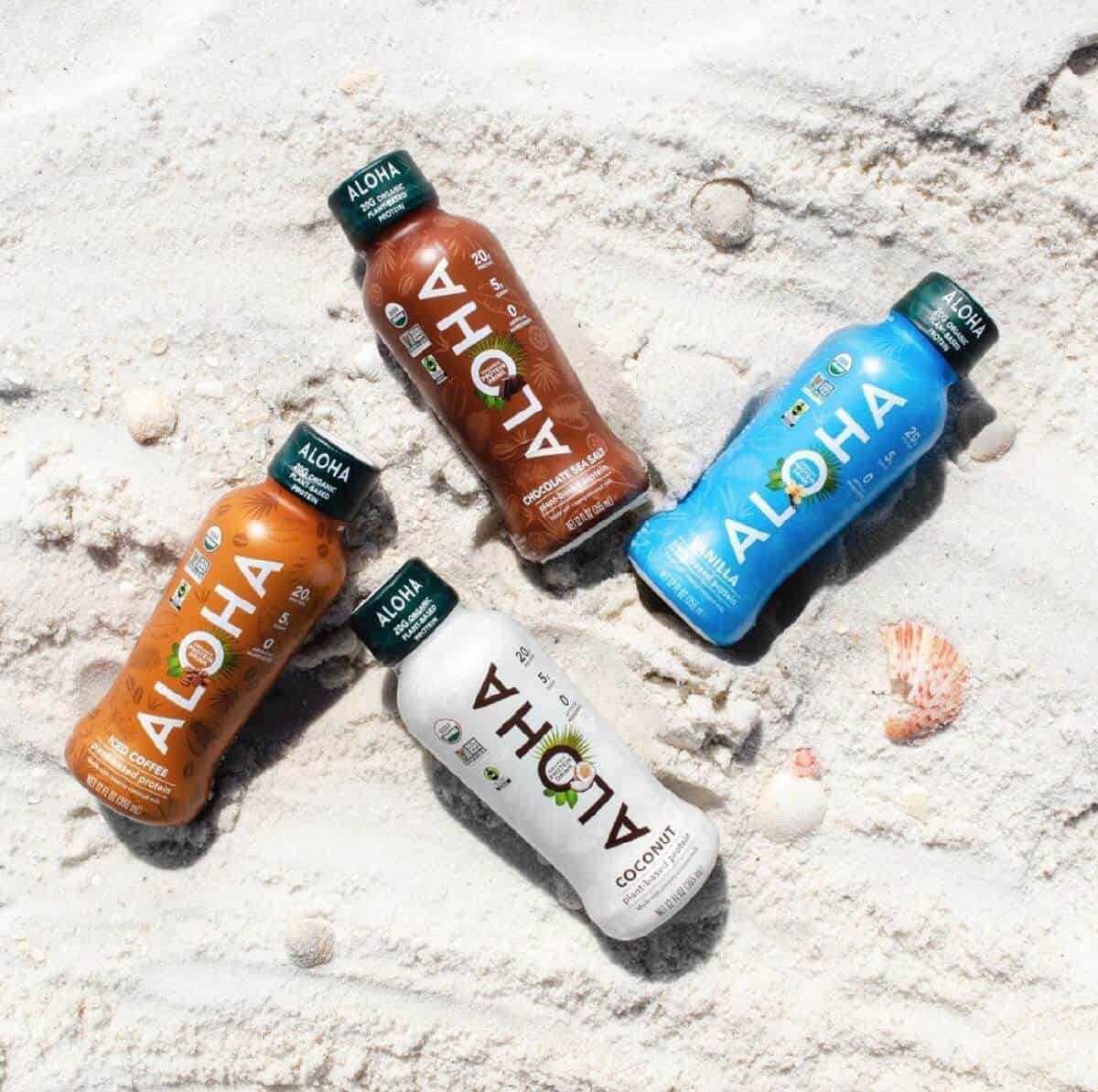 Four bottles of Aloha Protein Shakes in blue, white, orange, and brown colors laying in the sand on the beach.