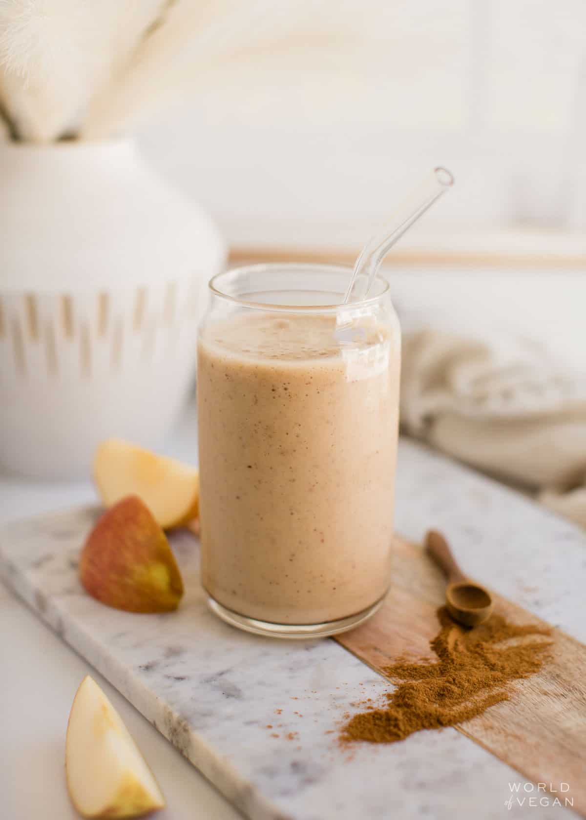A glass filled with apple pie banana smoothie with sliced apples and ground cinnamon next to the glass.