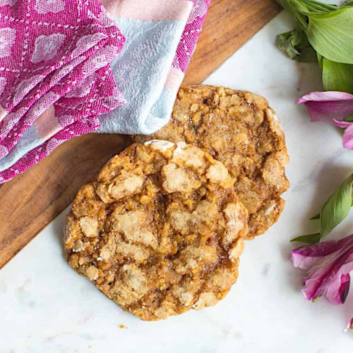 Two vegan oatmeal cookies on a marble serving platter next to fresh flowers and a mug of coffee.