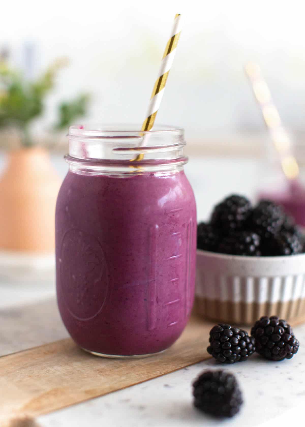 Blackberry smoothie in a mason jar with a straw, beside a bowl of fresh blackberries.