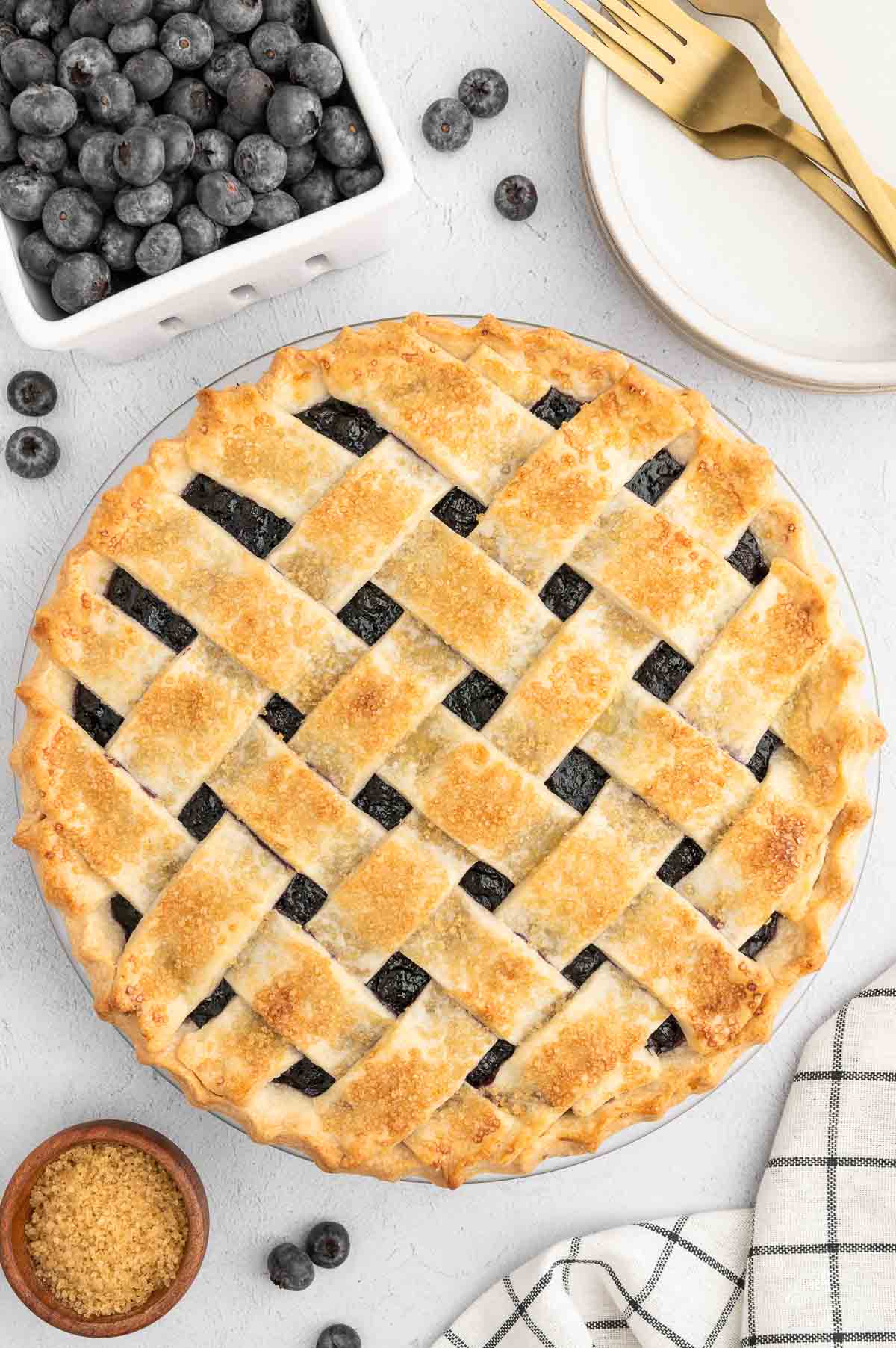 A whole vegan blueberry pie with a golden lattice crust, sitting next to a carton of blueberries.