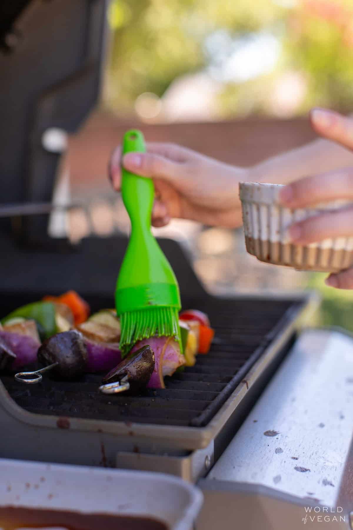 A hand brushing more marinade onto the tofu kebabs on the grill.