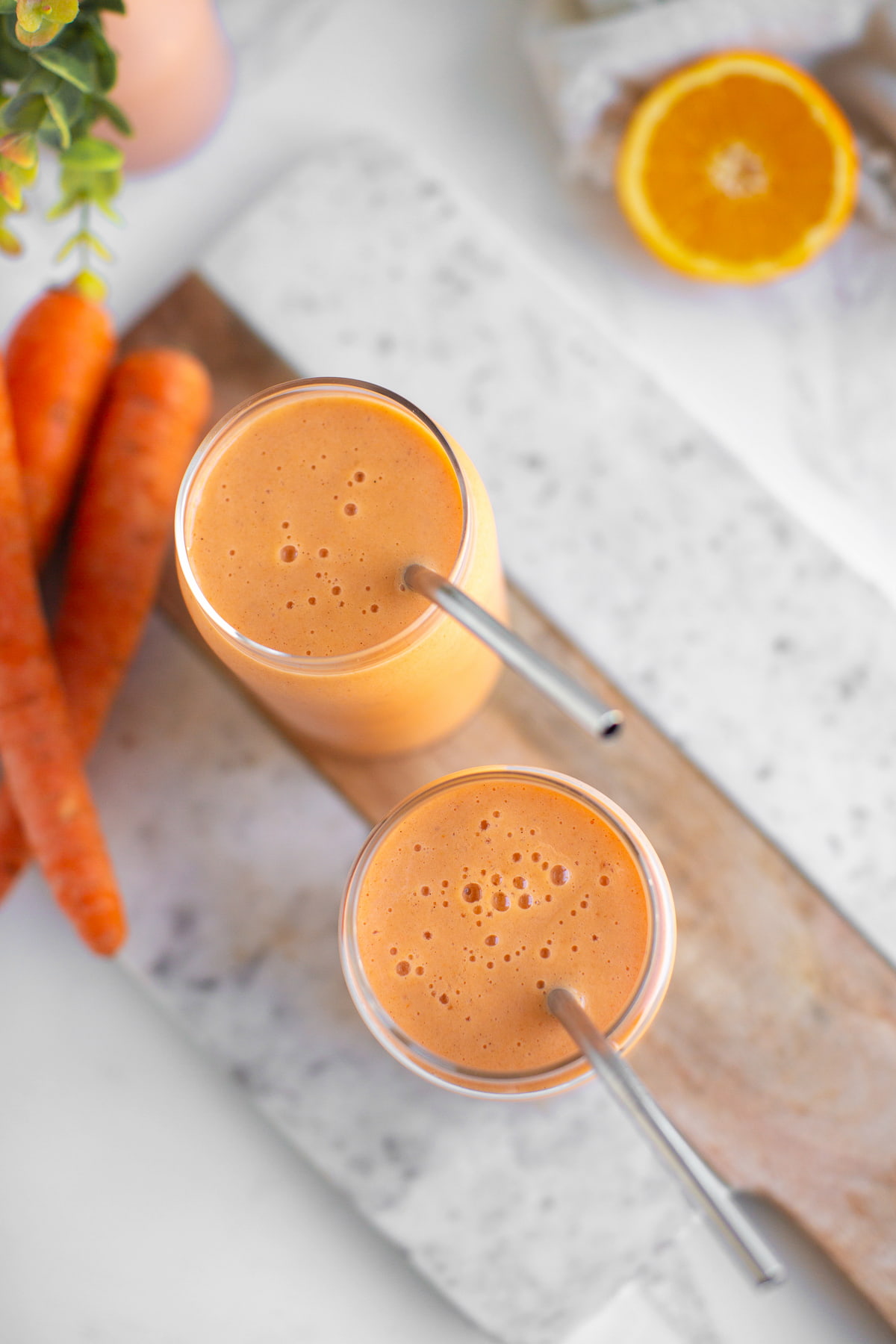 Two carrot smoothies in glass cups with metal straws next to a few carrots.