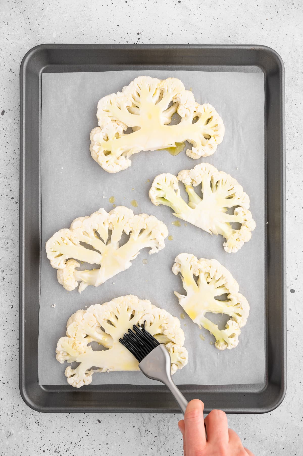 A basting brush applying oil to the cauliflower steaks on a baking tray.