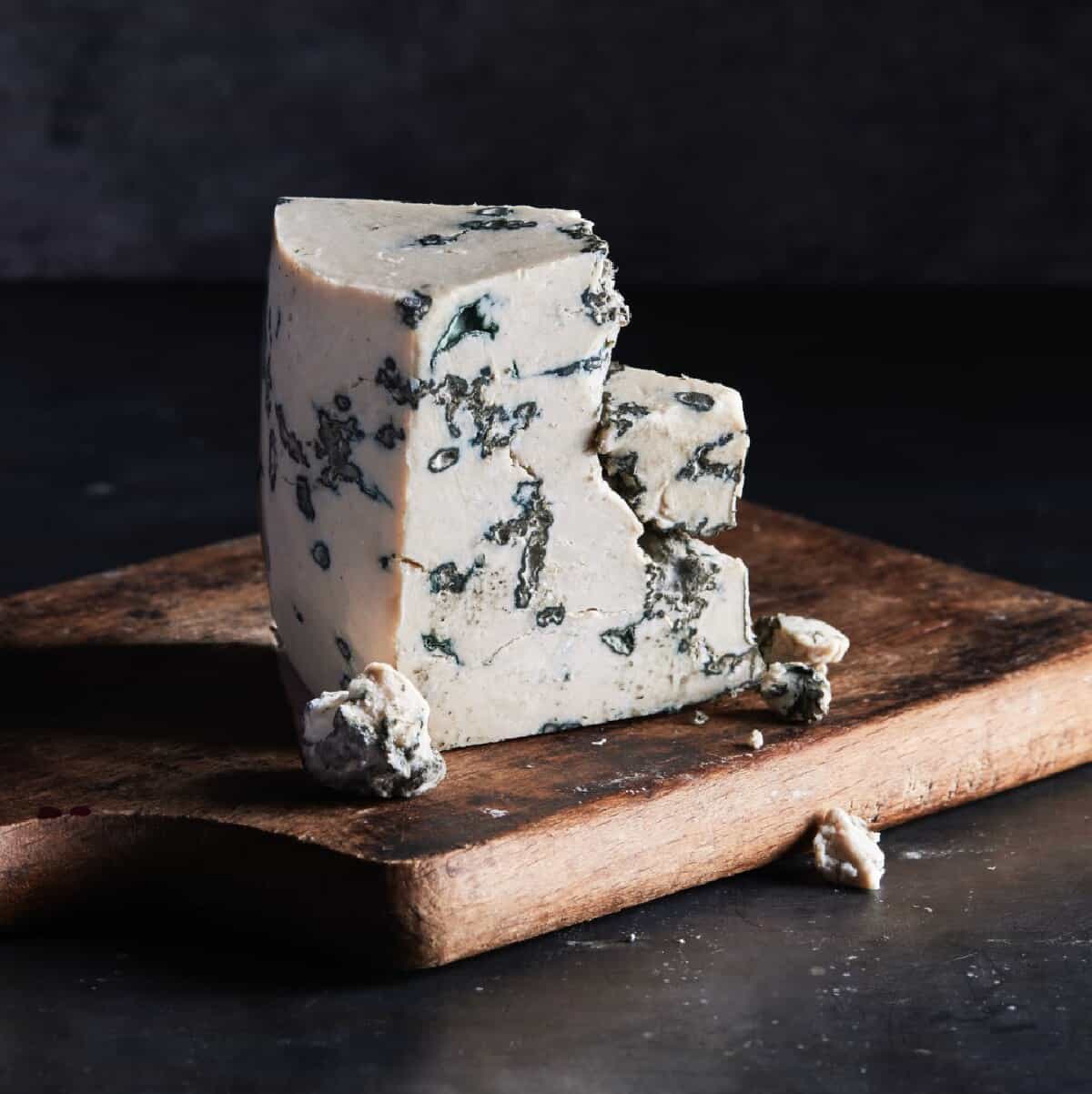 A wedge of Climax's dairy-free, vegan blue cheese on top of a wooden cutting board against a black background. 