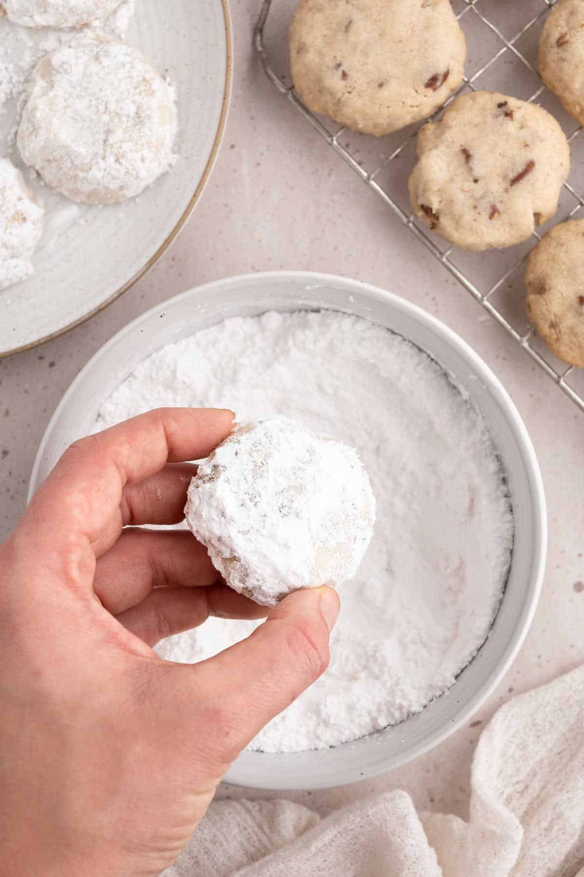 Hand holding pecan sandie cookie over bowl that has been dipped in powder sugar.