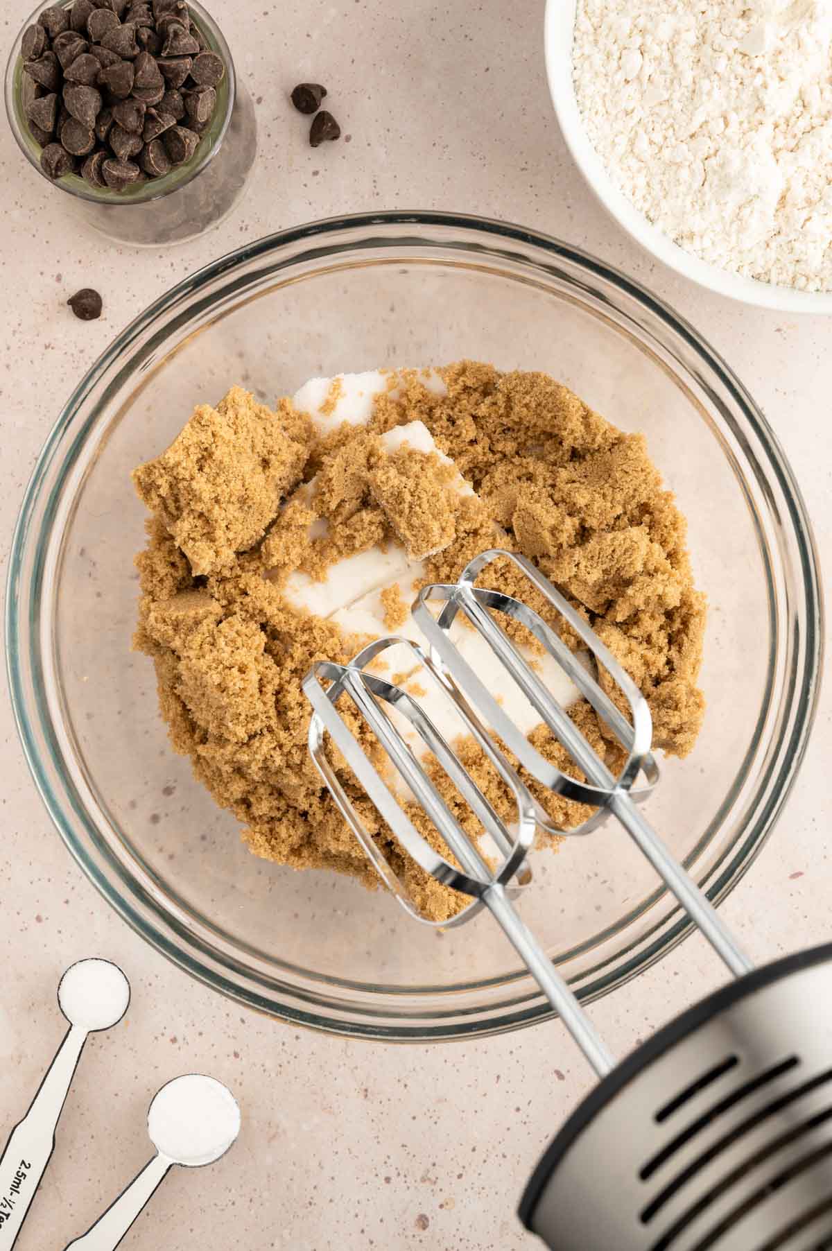 A glass bowl of cookie dough being mixed together with an electric mixer.