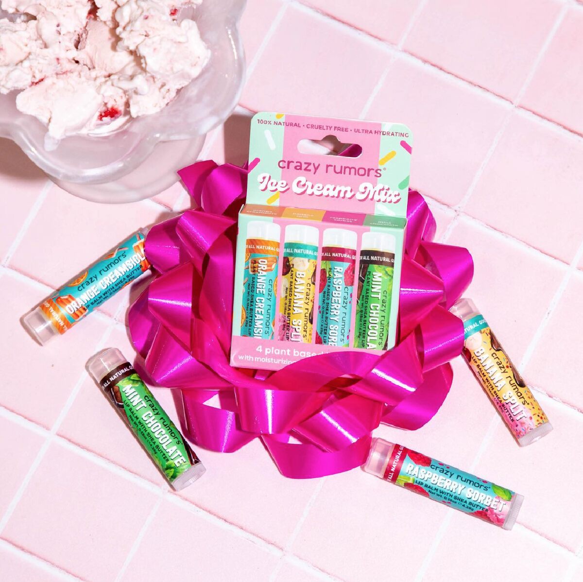 A colorful box of Crazy Rumors Ice Cream Mix vegan lip balms on top of a hot pink bow and next to a bowl of strawberry ice cream on a pink tile background. 