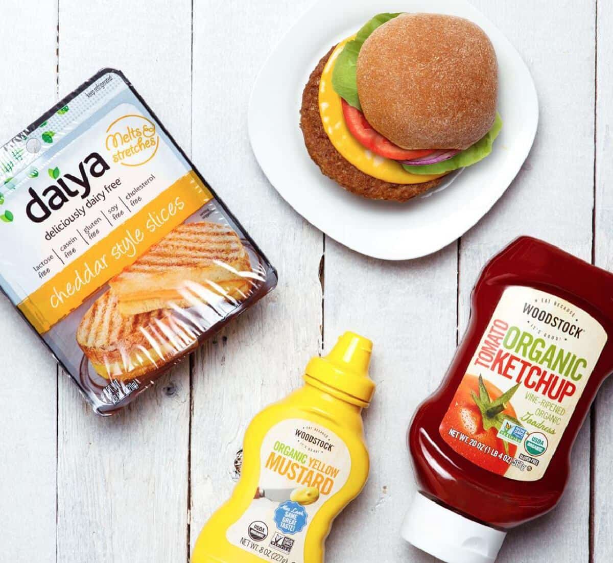 A package of Daiya vegan cheese slices, a bottle of ketchup, mustard, and a veggie burger on a plate against a white wooden background. 