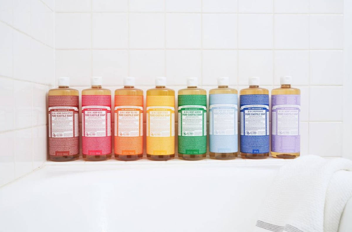 Eight bottles of Dr. Bronner castile All-One soap lined up in rainbow order against white bathroom tiles. 