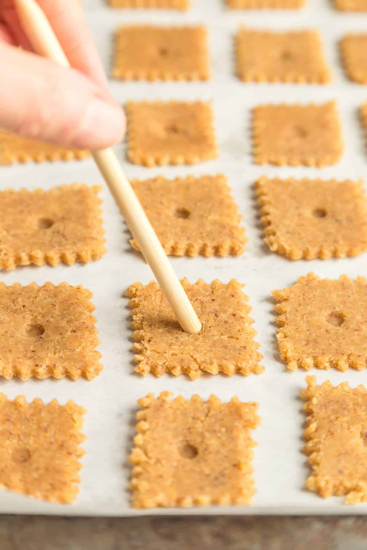 Almond flour dough scored into squares and placed on a baking sheet with parchment paper. A wooden stick is piercing a hole into each cracker.