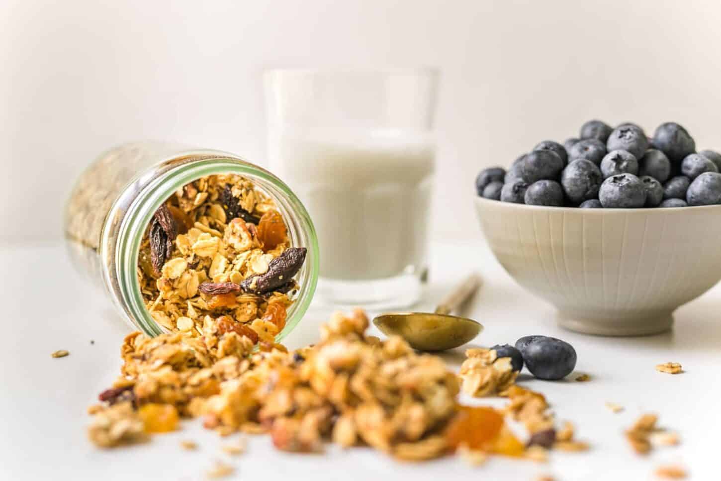 Mason jar filled with homemade granola spilled on white surface and a glass of plant milk in the background