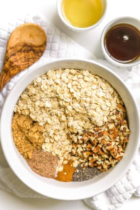 Large white bowl with homemade granola ingredients, a small bowl with maple syrup on the side