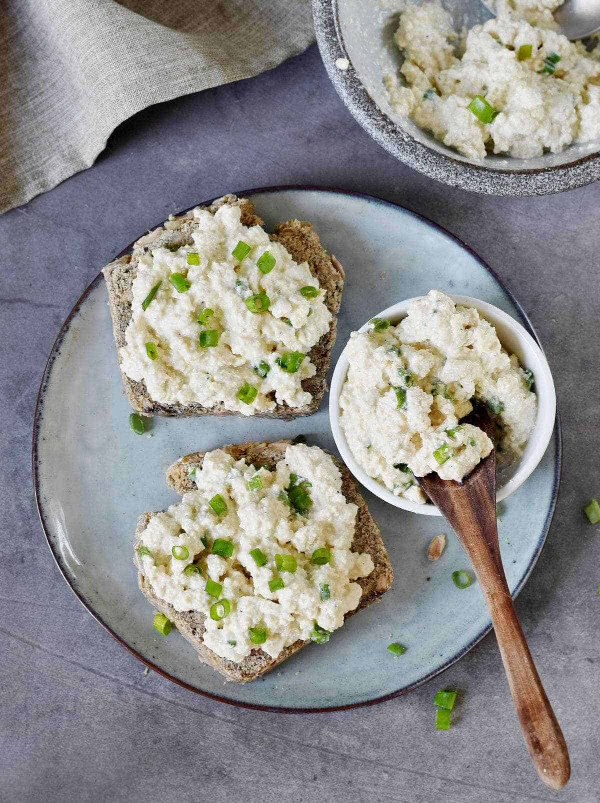 Two slices of bread covered with vegan cottage cheese and chives, as well as a small white bowl filled with cottage cheese and a wooden spoon on a gray stone circular plate on a gray countertop accented with a gray cloth napkin.