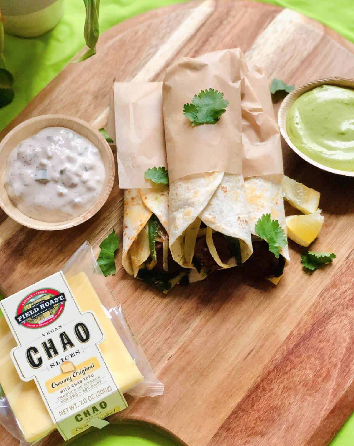 A wooden cutting board filled with rolled up tortillas, creamy dips, a lemon wedge, and a package of Field Roast vegan Chao cheese slices. 