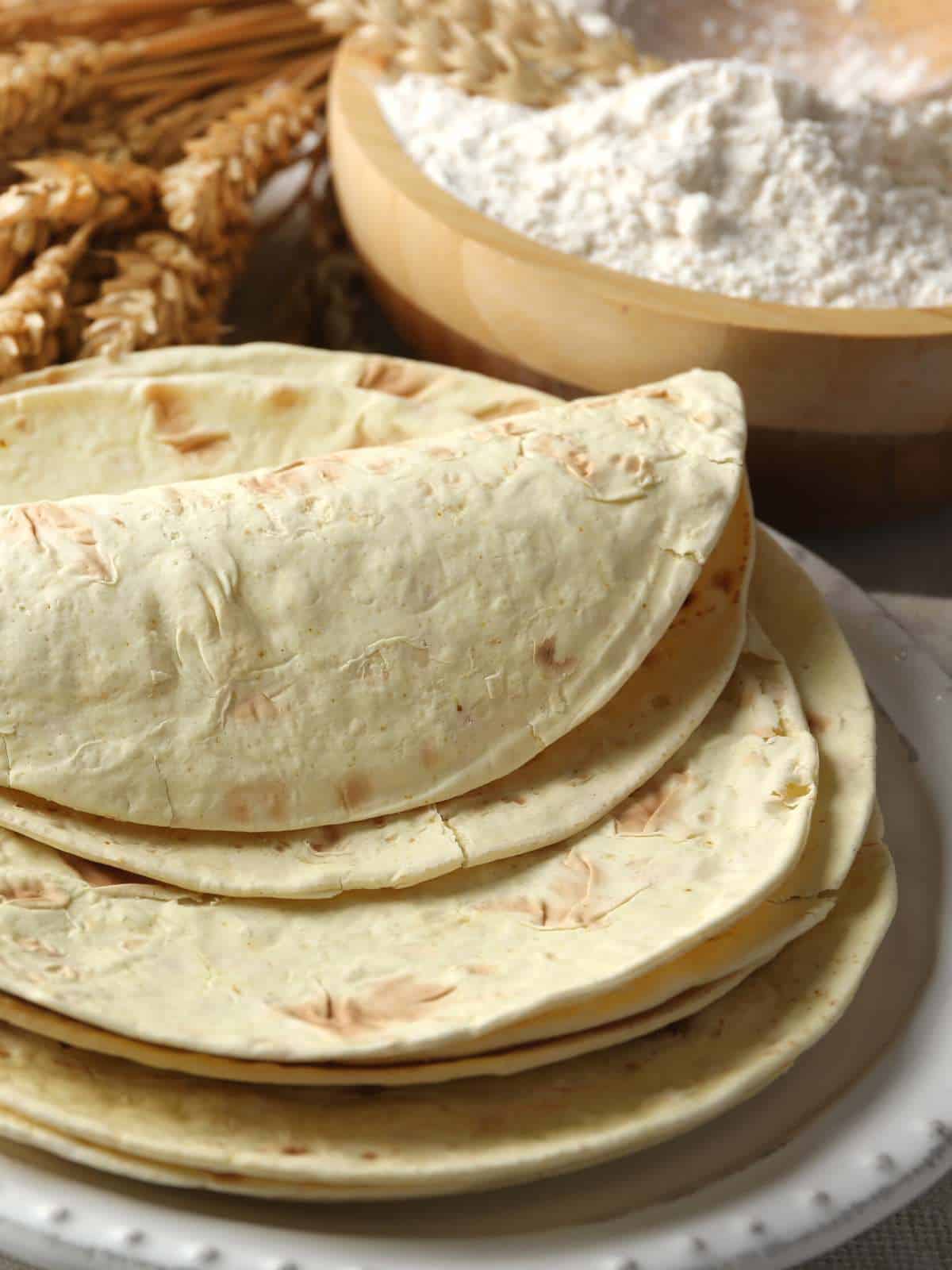 Whole wheat flour tortillas in a stack with one folded in half and a bowl of whole wheat flour behind it.