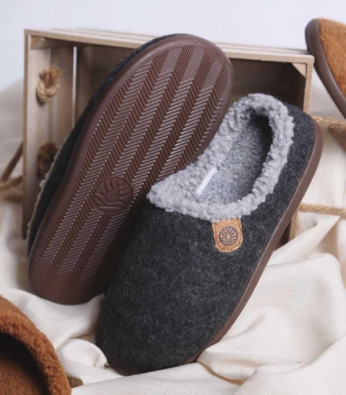A pair of vegan wool and vegan fur mule slippers in dark gray leaning upright against a small wooden crate.