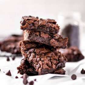 Pile of Fudgy Vegan Brownie Squares on a white surface with chocolate chips sprinkled around.