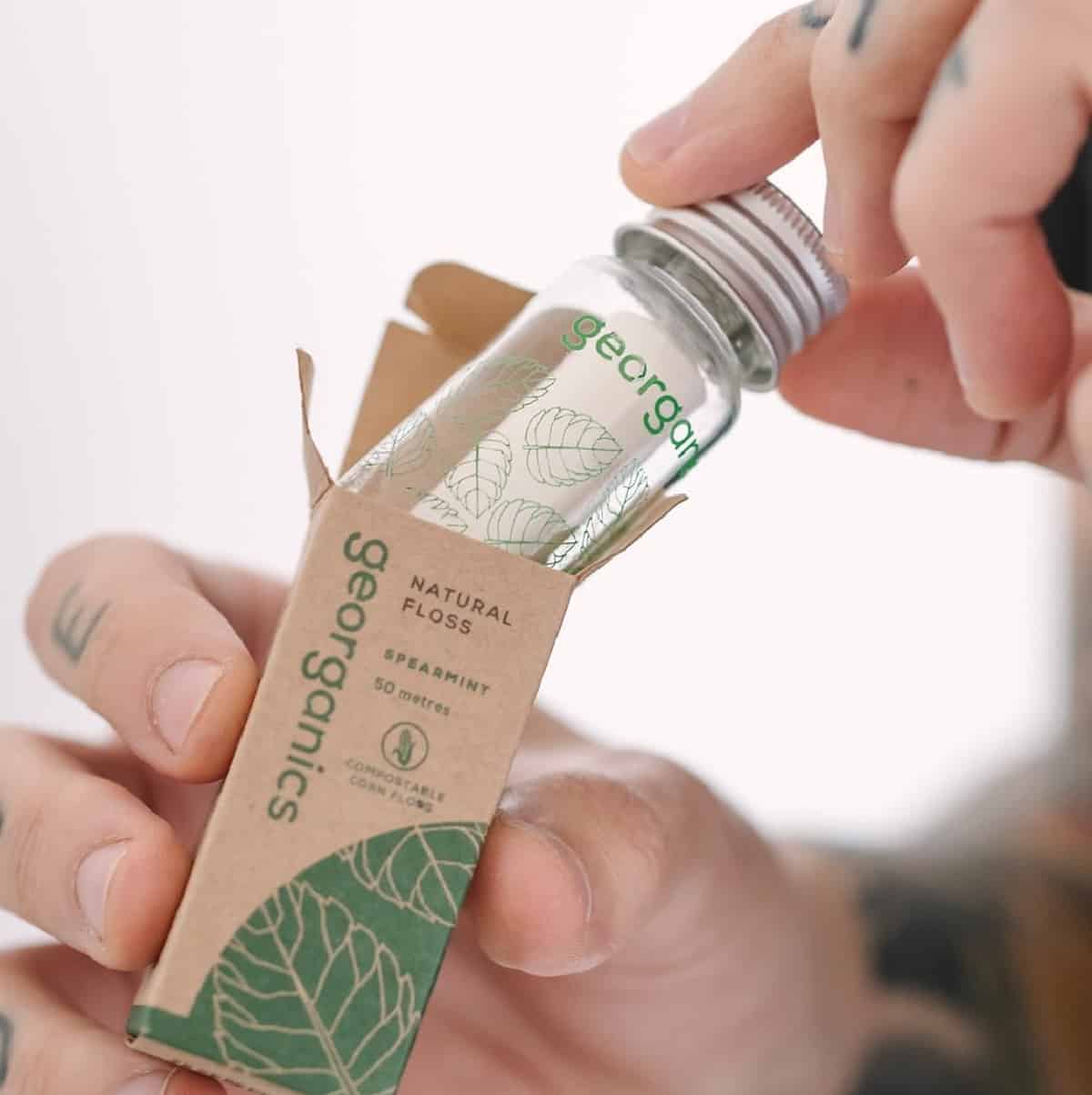 Hands pulling out a glass bottle of Georganics vegan floss out of a small cardboard box. 