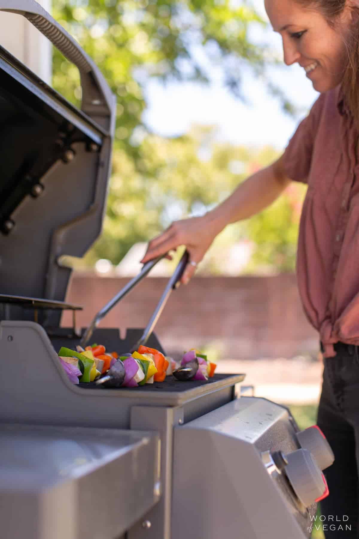 A hand holding tongs, flipping tofu kebabs over on the grill.