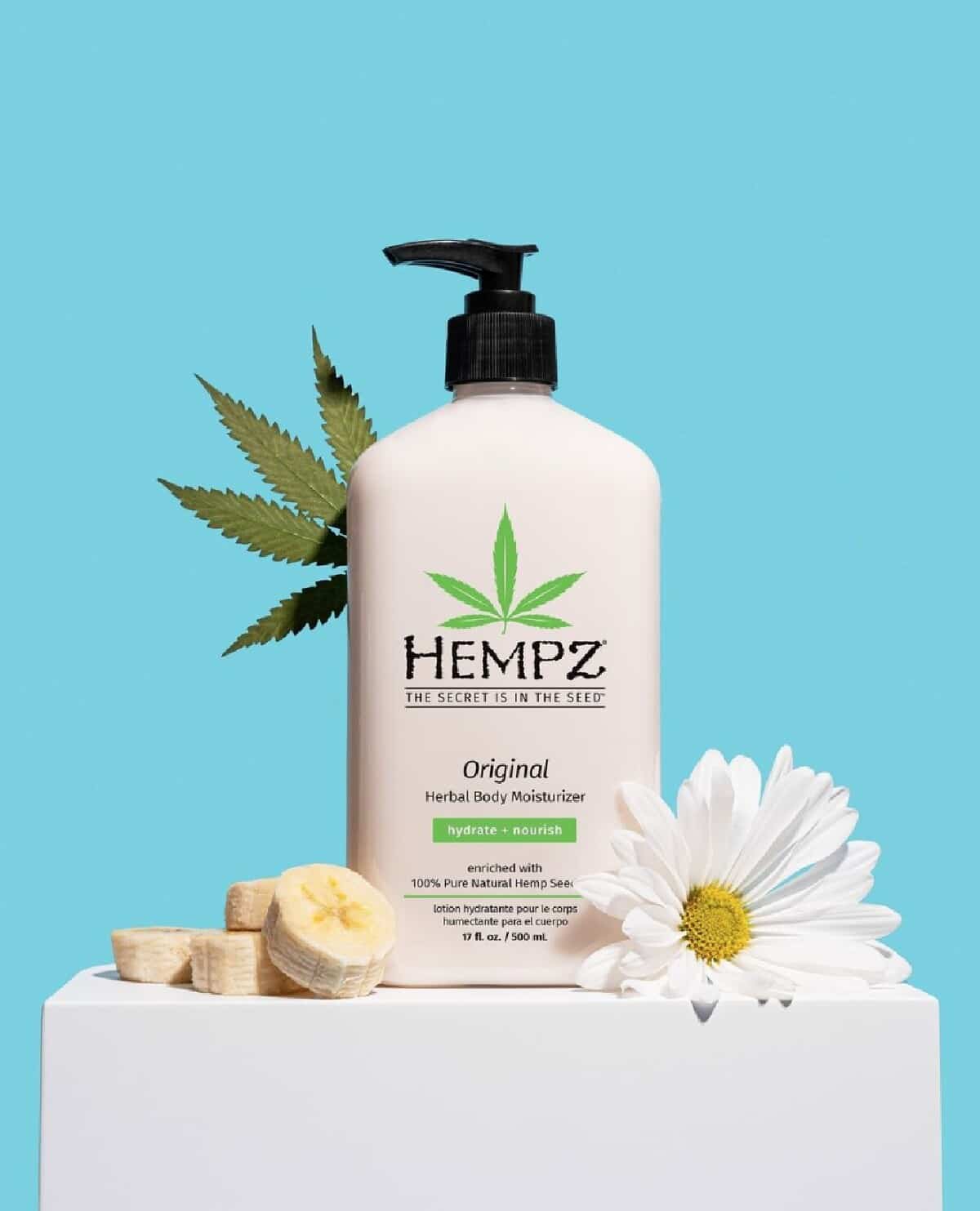 A white bottle of Hempz body lotion with a black pump top on top a white box decorated with bananas, daisies, and a hemp leaf on a blue background. 