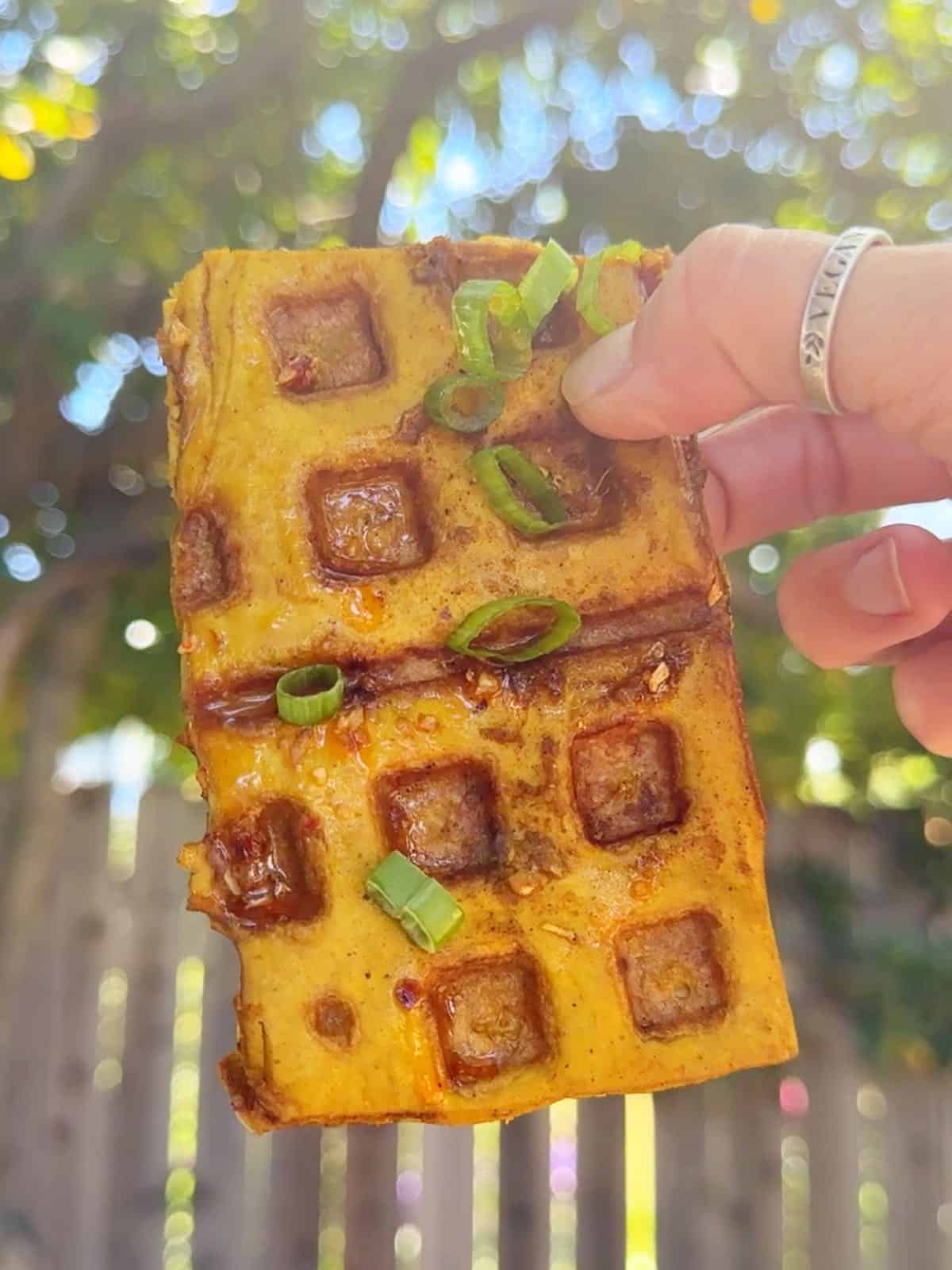 Vegan woman holding out grilled tofu waffles made in a waffle iron with and outdoor scene in the back. 