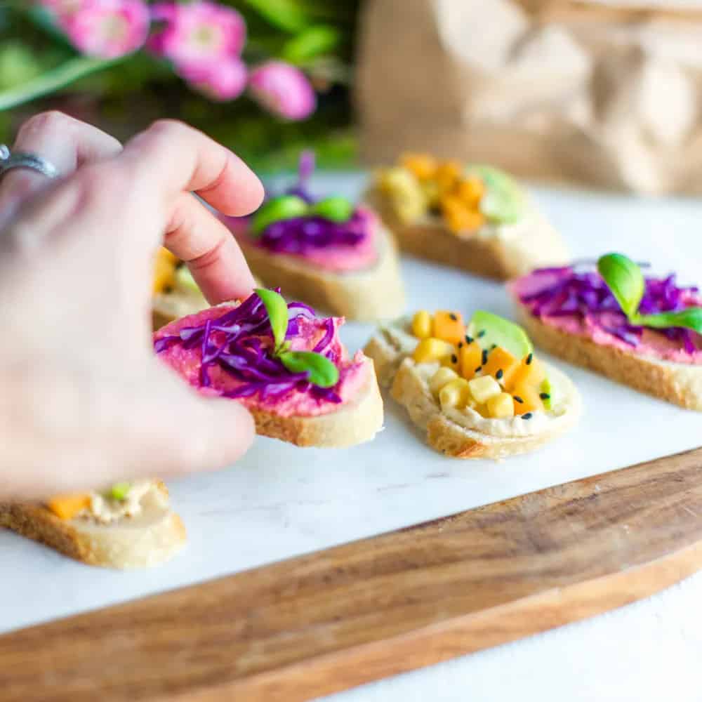 white hand holding crostini topped with beet hummus, cabbage, and basil garnish. there's crostini with white hummus, corn, and orange squares in the background too