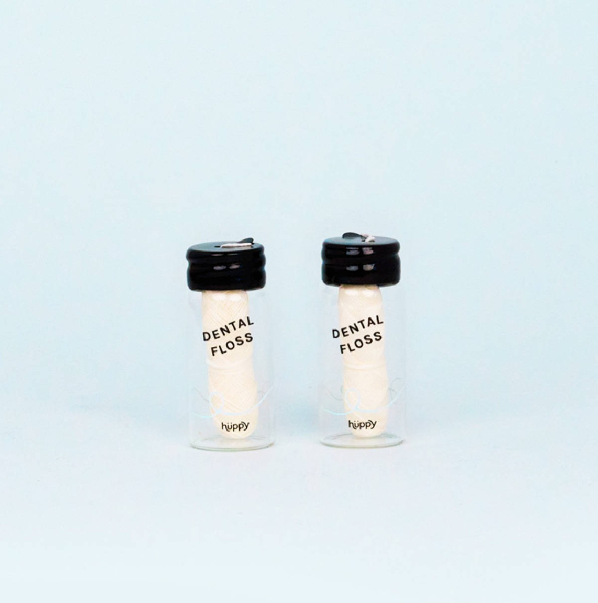 Two glass containers of Huppy floss with black lids against a pale blue background.