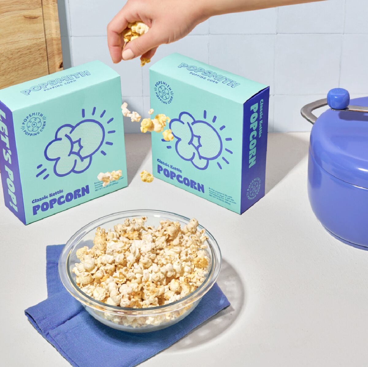 Two blue and purple boxes of Popsmith popcorn next to a purple popcorn popper and a bowl of prepared popcorn on blue napkins. 