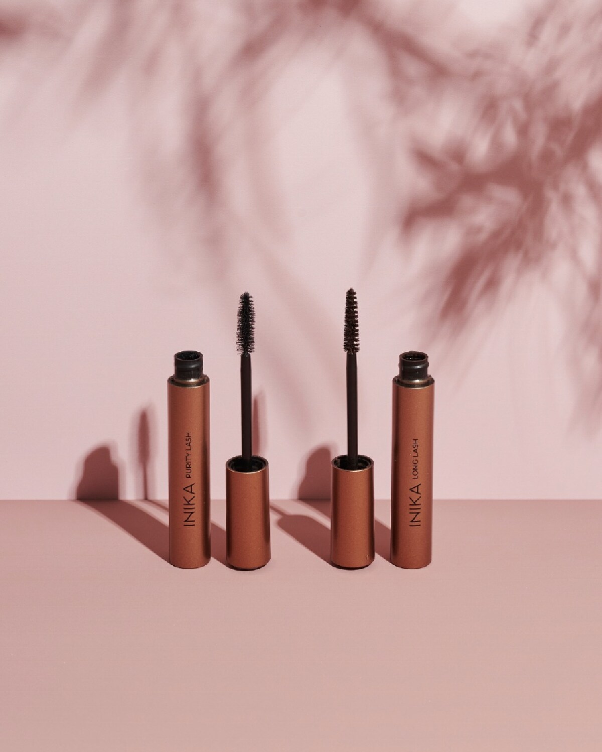Two copper colored opened tubes of Inika Organics mascara next to black mascara wands against a shadowed pink backround. 