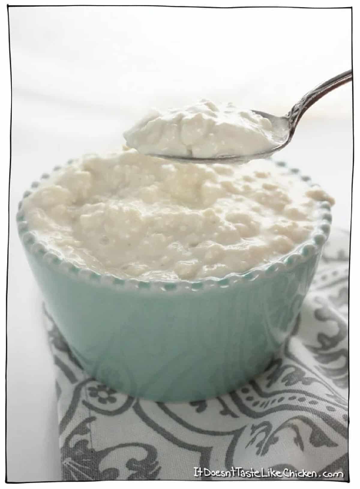 A pale turquoise bowl filled with vegan cottage cheese and a spoonful held above the bowl against a white background and displayed on a white and gray patterned towel.