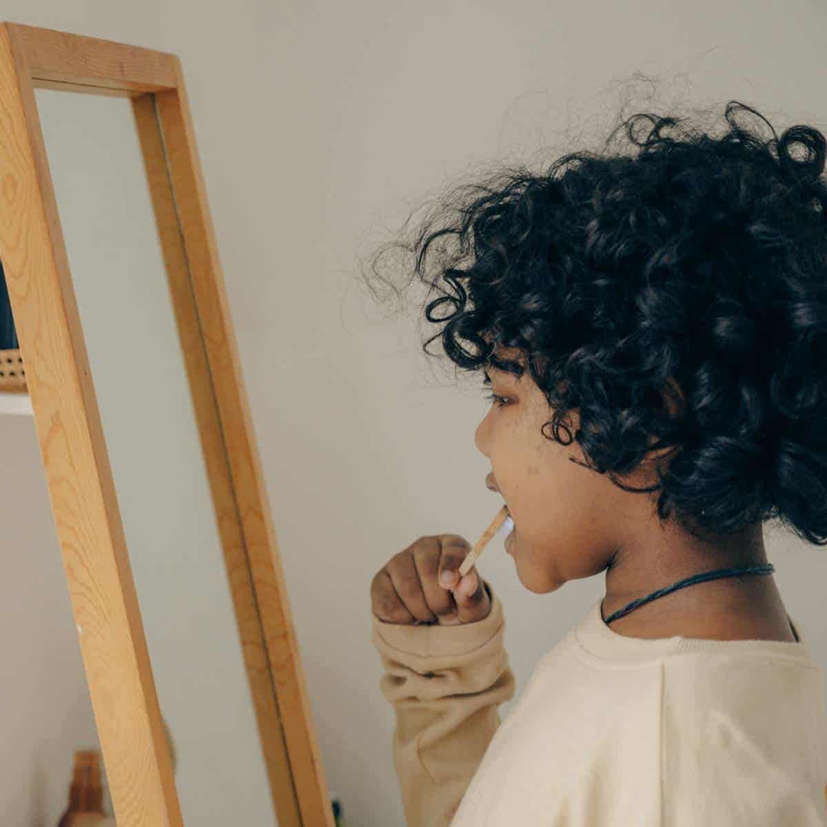 Toddler child with black curly hair standing in front of a mirror brushing his teeth with a wood toothbrush. 