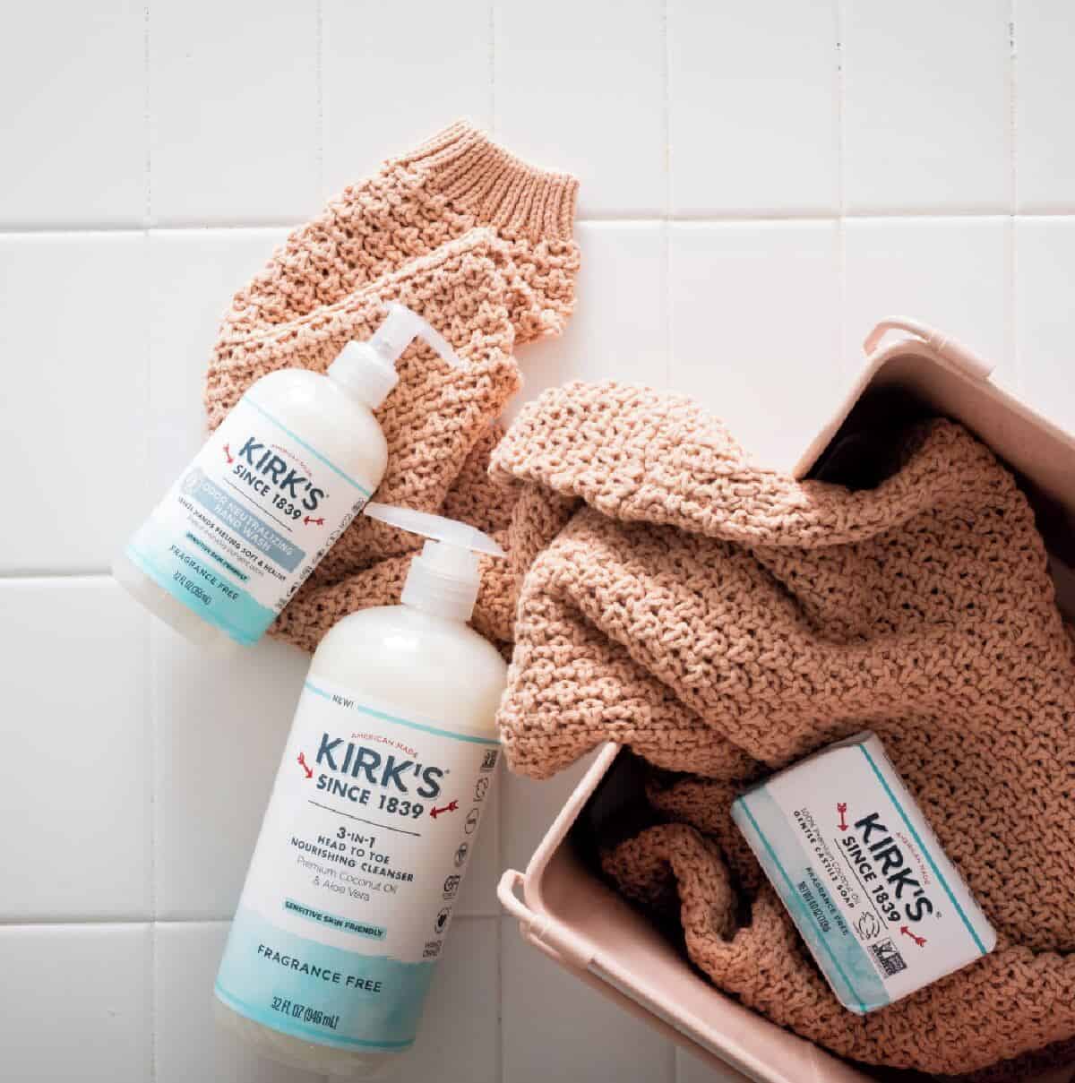 Two Kirk's pump fragrance-free pump bottles and one bar of soap laying on a hand knit light brown sweater on a white tile background.