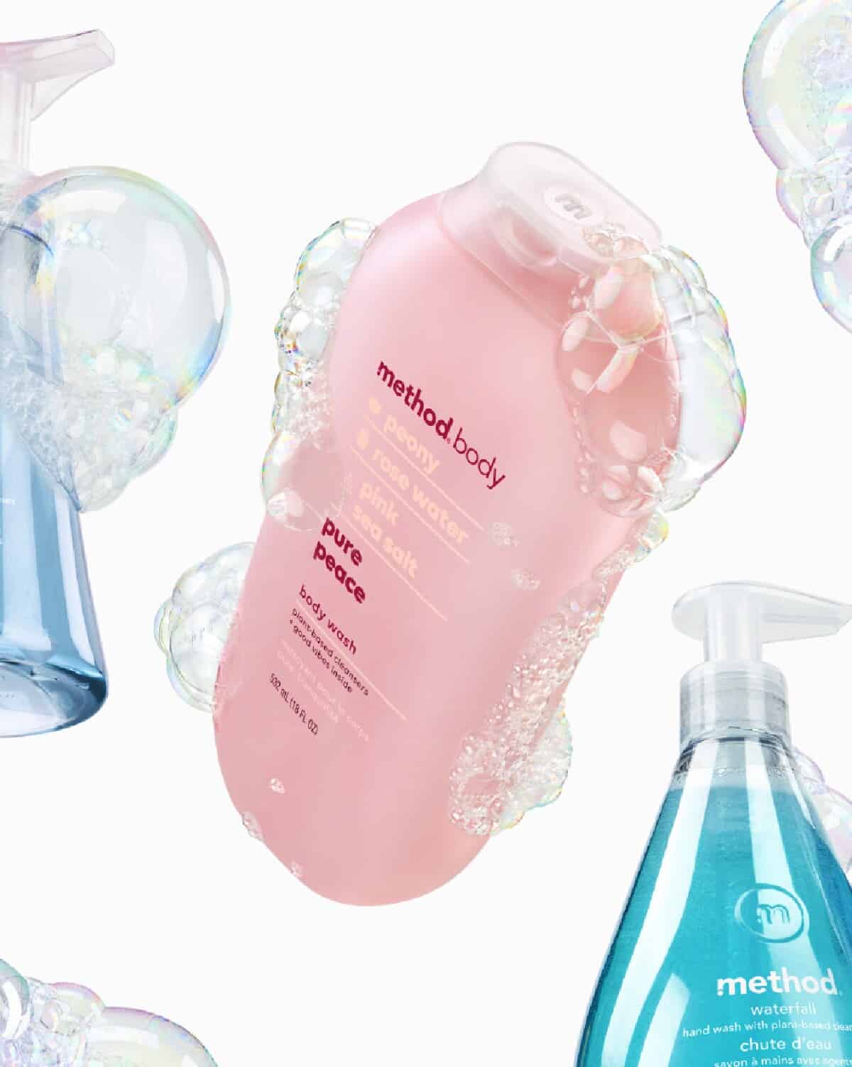 A pink container of Pure Peace Body Wash by Method Body against a white background with bubbles and clear containers of hand wash. 