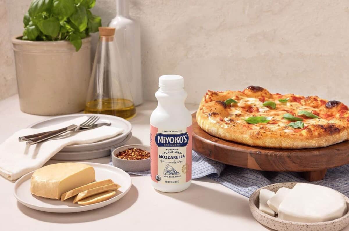 A white and pink bottle of Miyoko's Creamery plant milk pourable mozzarella on a table with vegan pizza, plates and silverware, and rounds of vegan cheese on a white tabletop.