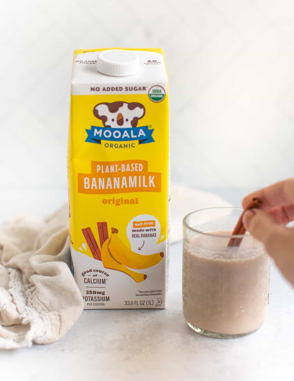 Glass of plant-based milk next to a carton of Mooala banana milk with a cinnamon stick. 