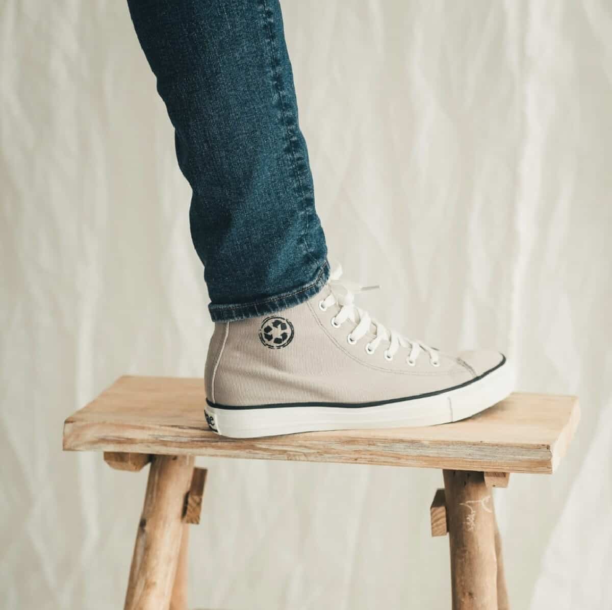 A beige and white high top vegan sneaker on a person with blue jeans leaning on a wooden stool with a white background.