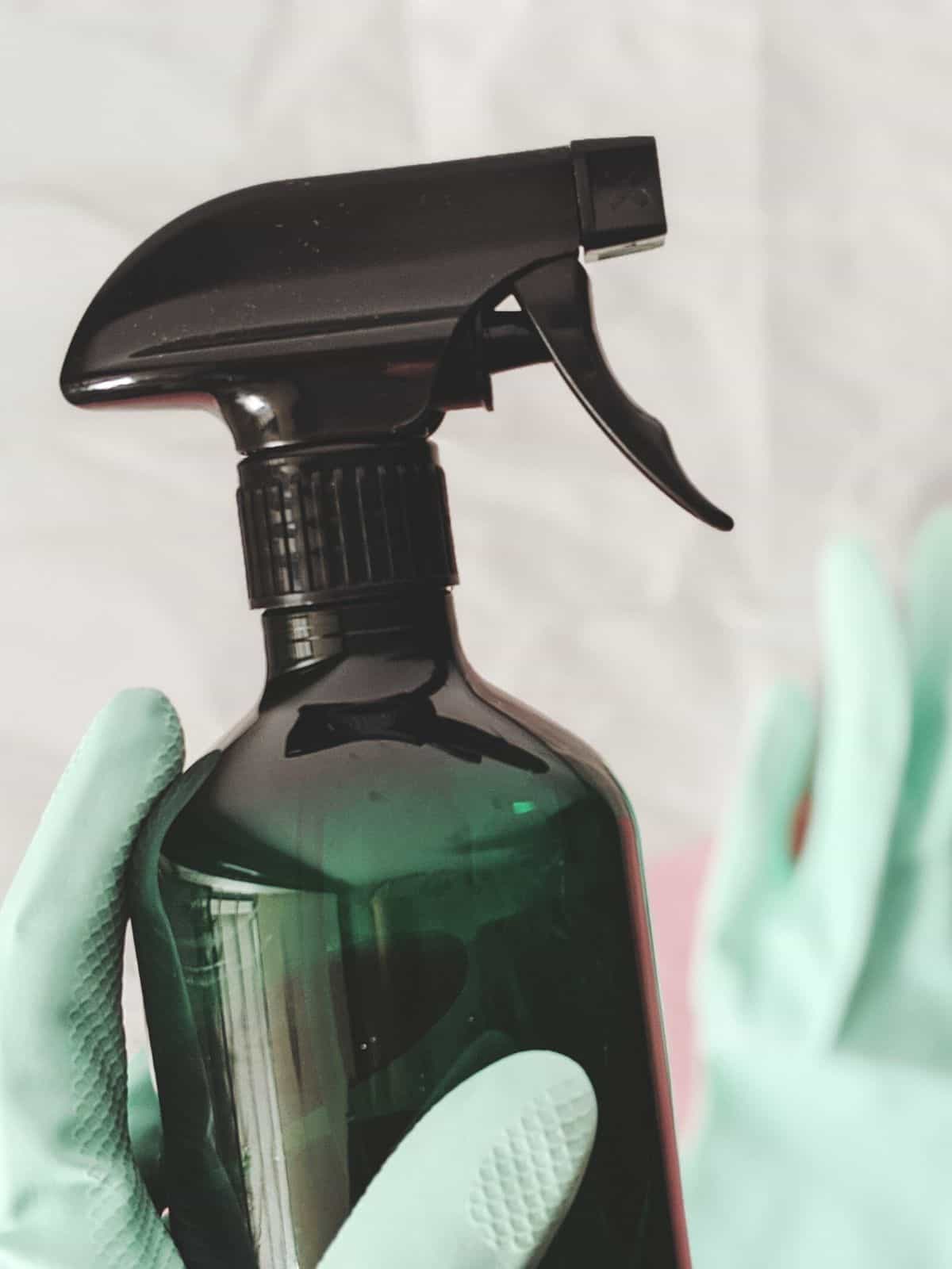 Hand with a green rubber glove on holding a glass spray bottle of natural homemade cleaner.