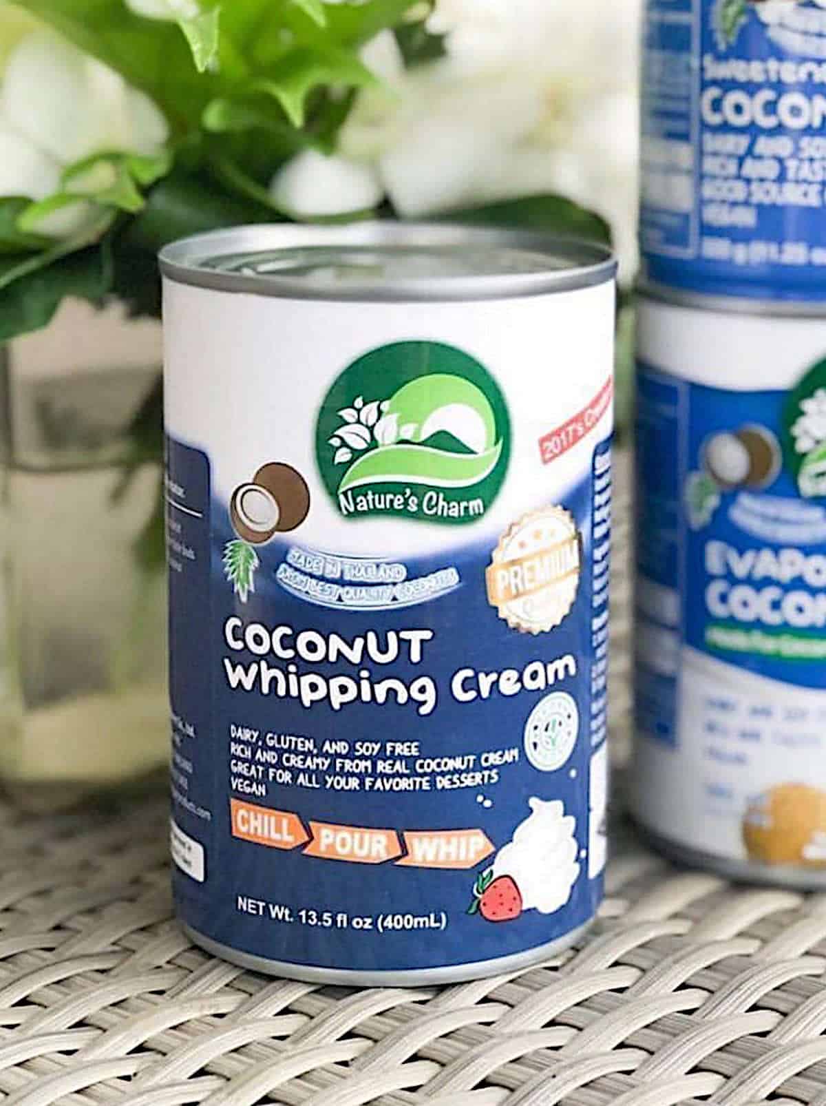 A can of Nature's Charm Coconut Whipping Cream on a weaved mat with flowers in the background.