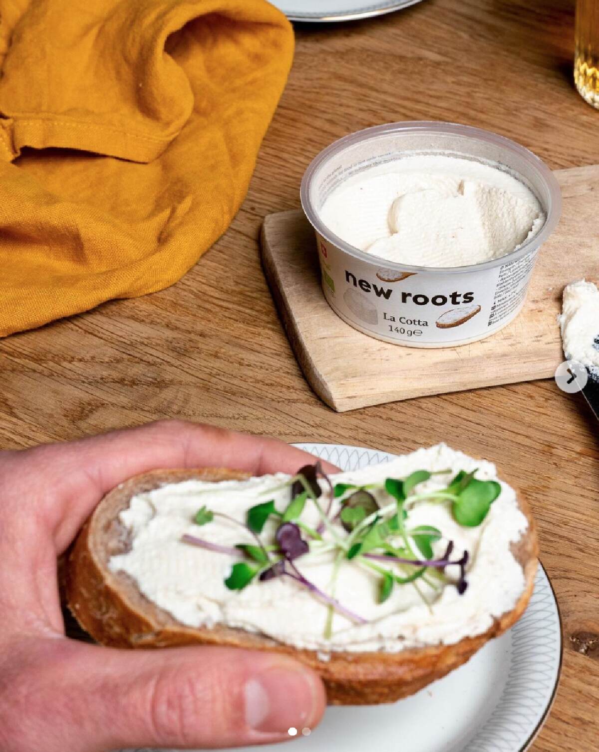 A hand holding a slice of bread covered in New Roots La Cotta cheese with sprouts. A plastic container of New Roots cheese is on a wooden cutting board nearby and an orange napkin against a wooden table background. 