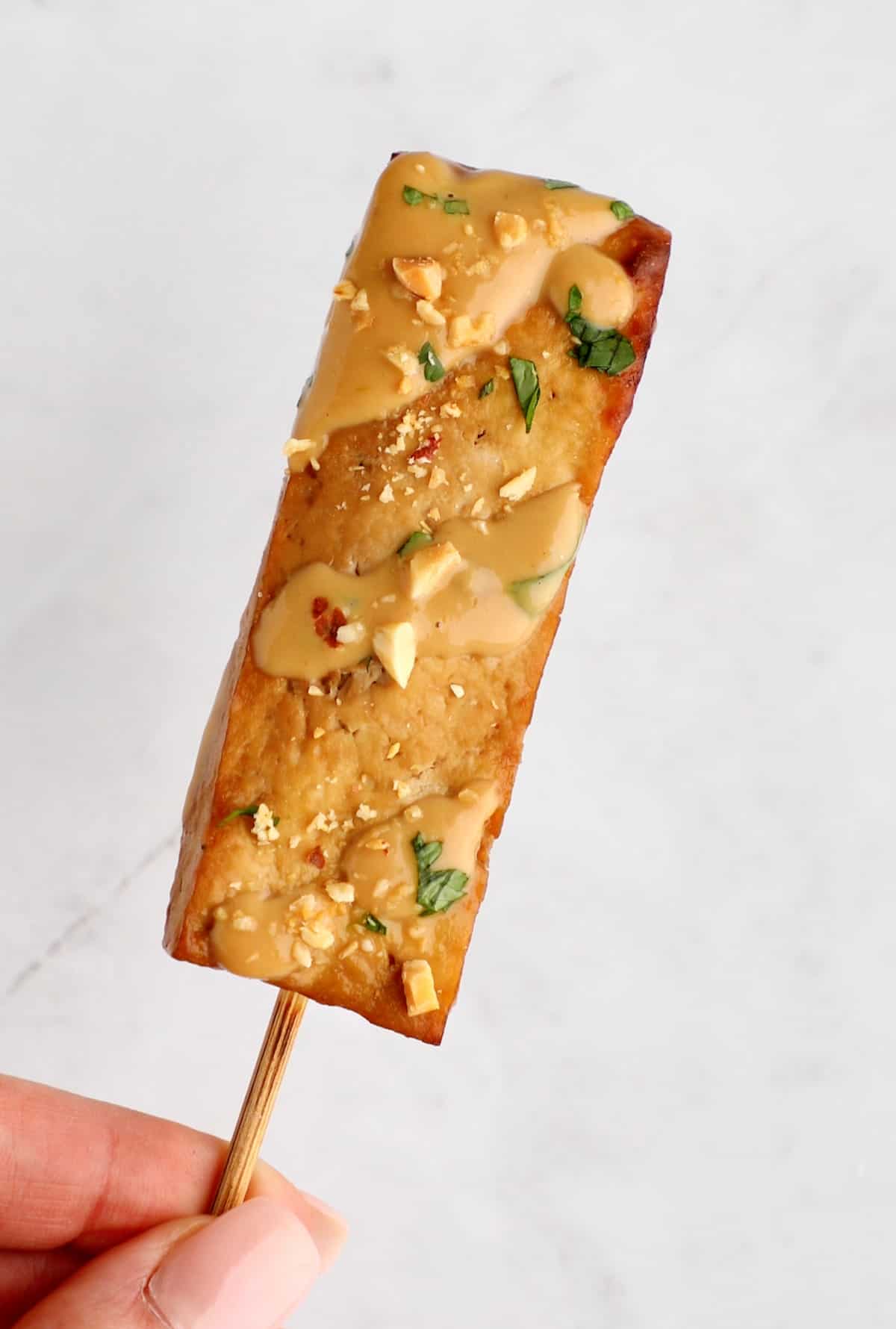 Hand holding one tofu satay skewer that's been drizzle with peanut dressing.