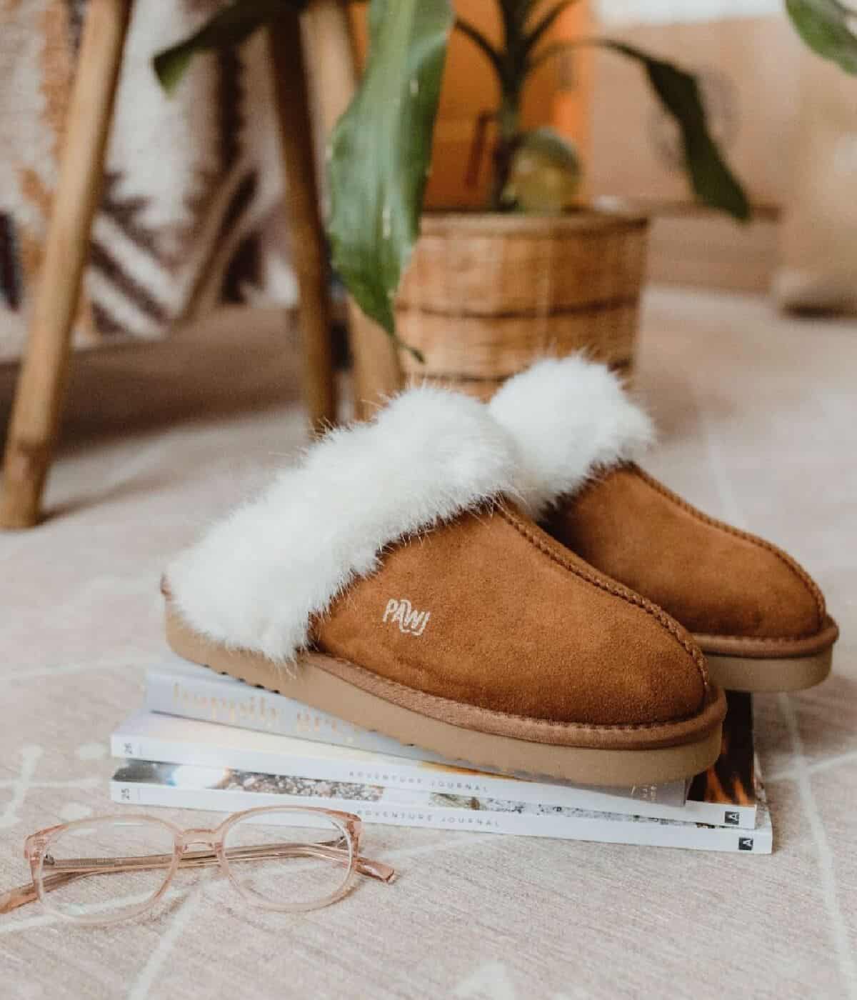 A pair of chestnut colored faux-fur lined slippers on top of a small pile of books next to a pair of pale pink eye glasses on the floor.