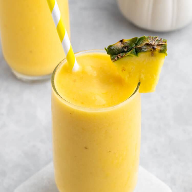 Pineapple mango smoothie in a glass with a straw and a slice of pineapple on the rim.