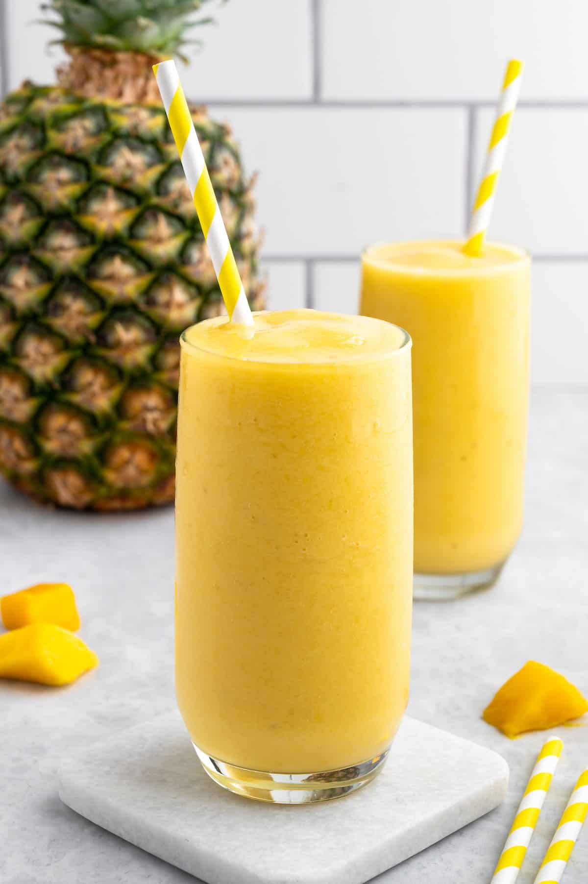 Two glass of mango pineapple smoothie with straws and a whole pineapple in the background.