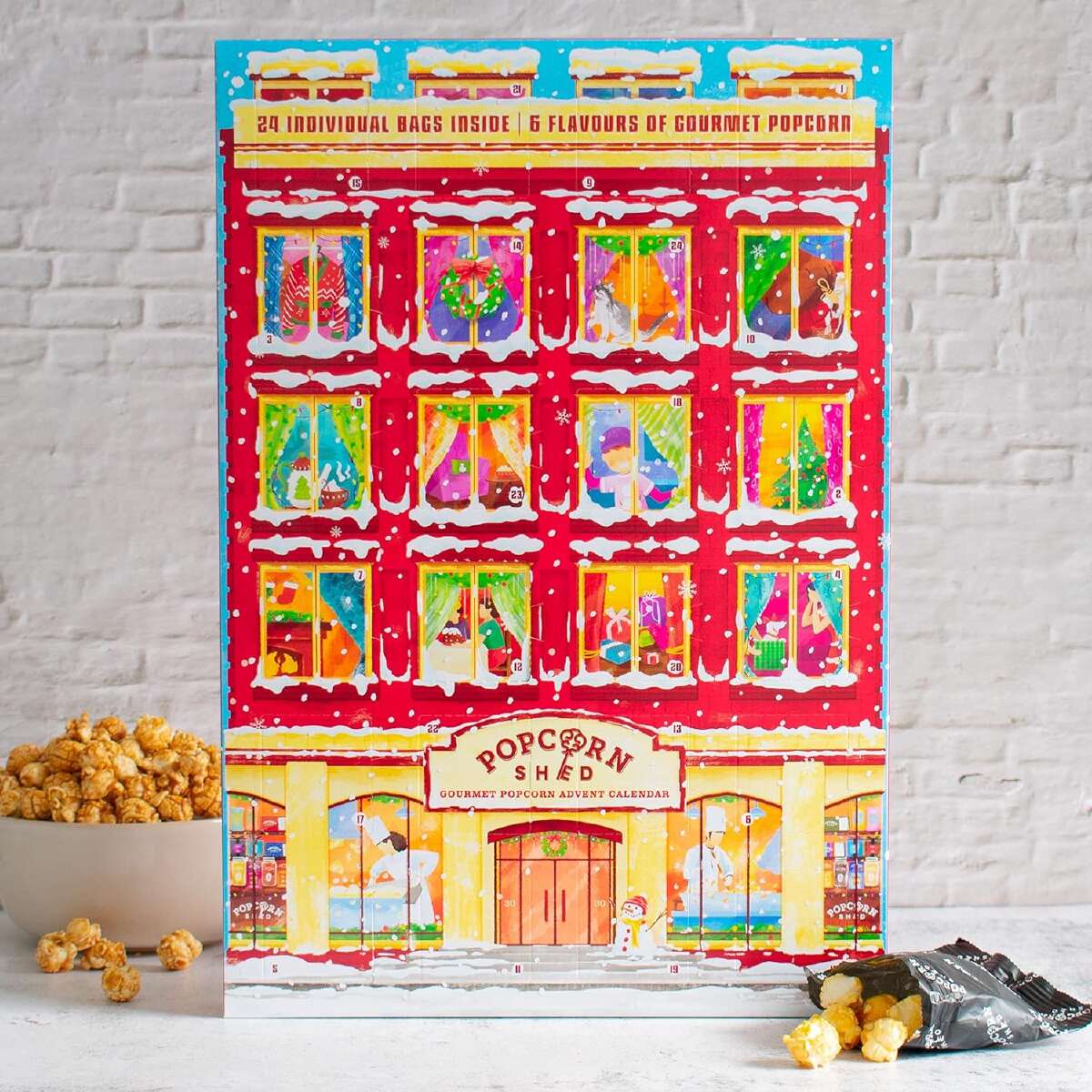 A popcorn advent calendar shaped like a large, fancy Christmas townhouse against a white brick wall background and accessorized with a bowl and opened packages of popcorn.