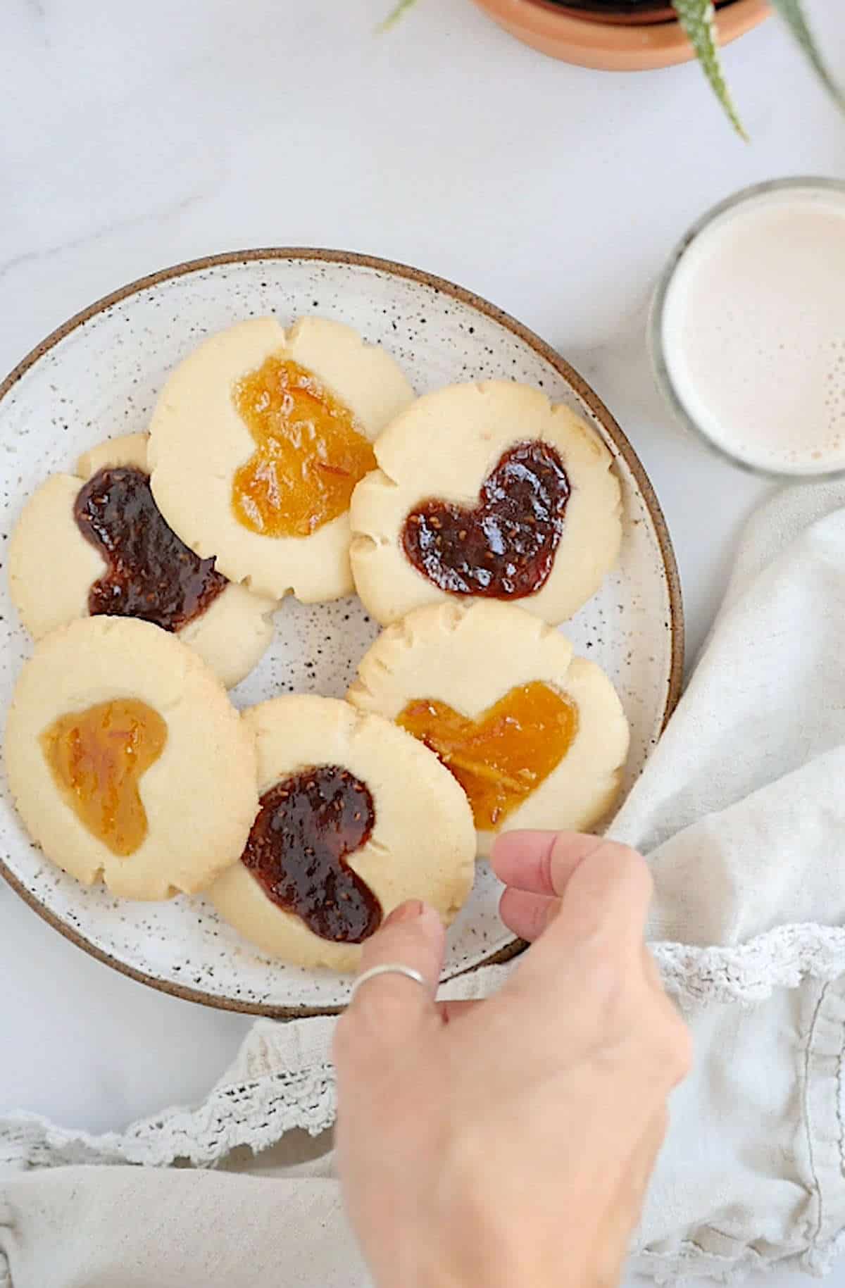 Vegan thumbprint cookies on a plate with a cloth napkin and a glass of non-dairy milk beside it and a hand reaching for a cookie.