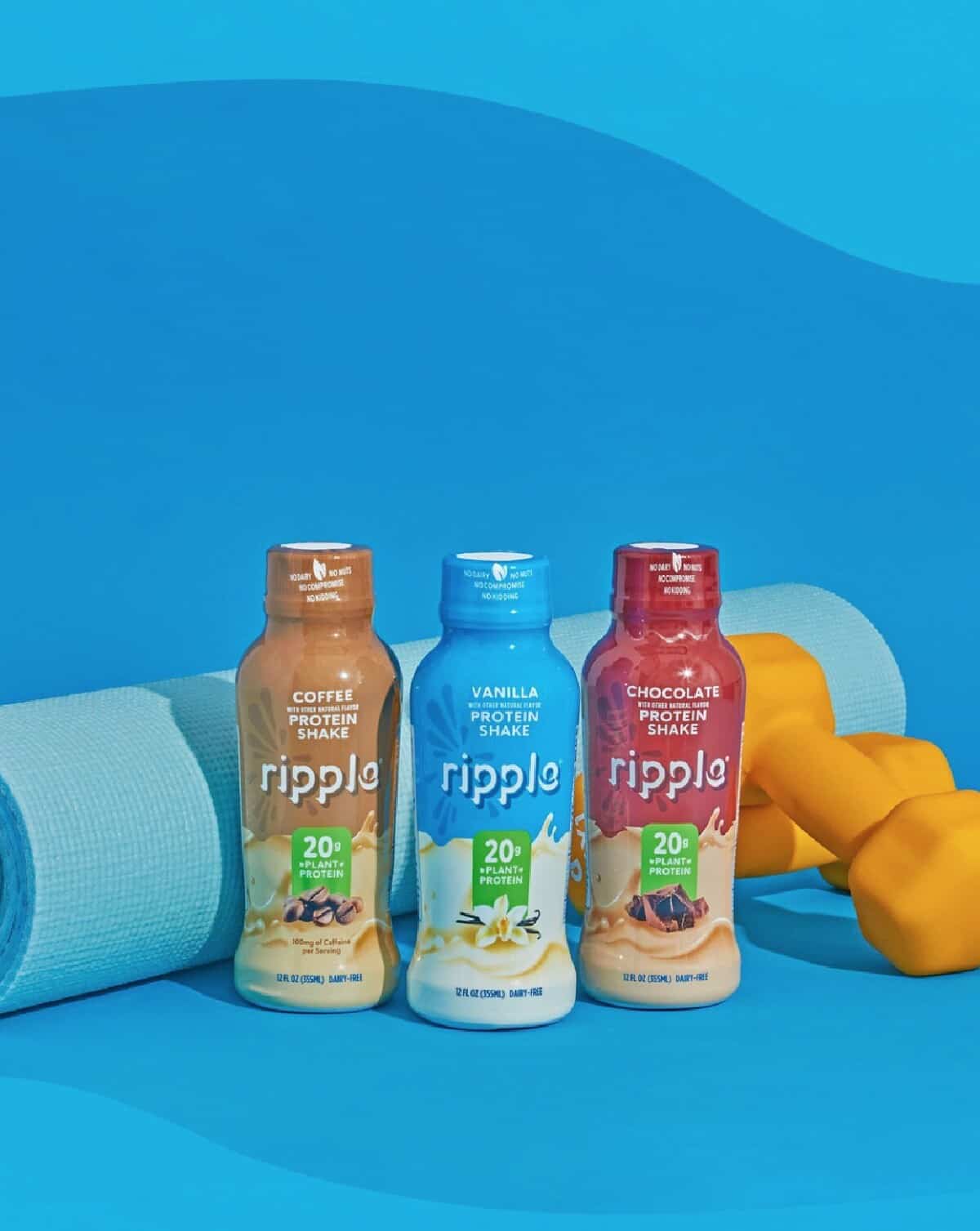 Three Ripple plant-based protein shakes in coffee, vanilla and chocolate standing against a yoga mat and set of weights on a blue background.
