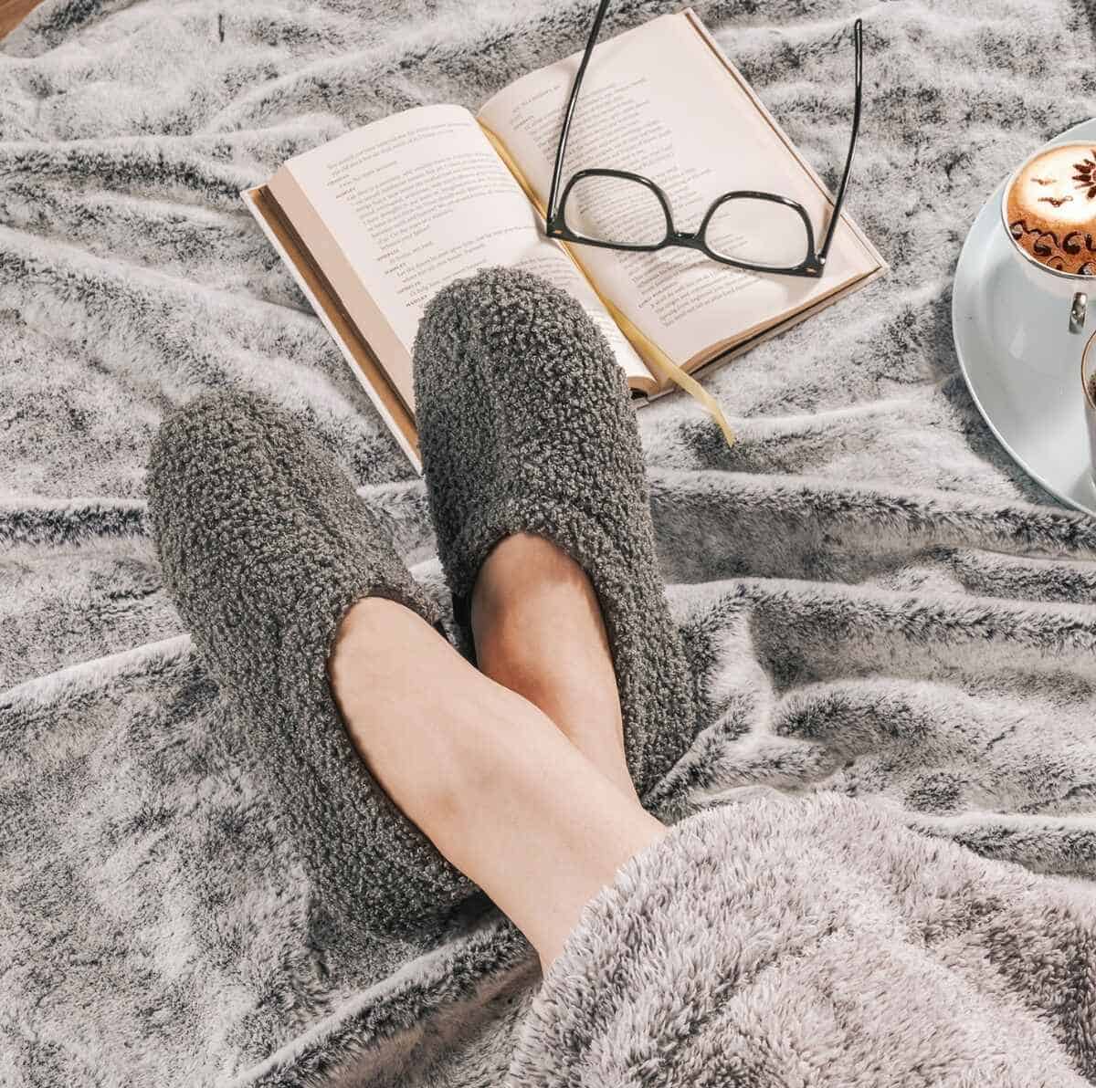 A dark gray pair of Rock Dove's indoor fuzzy fleece slippers on a woman's crossed feet surrounded by an open book with glasses and a luxurious gray blanket. 