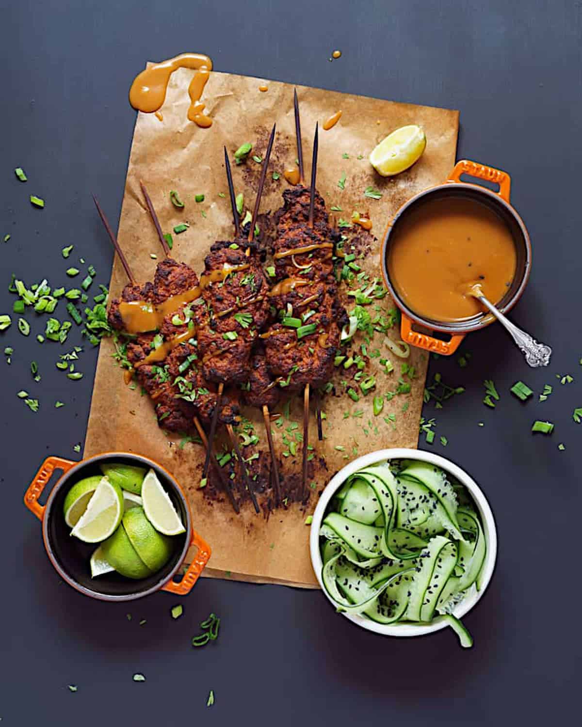 Vegan seitan kebabs on parchment paper, next to peanut sauce, lime wedges, and cucumber slices.